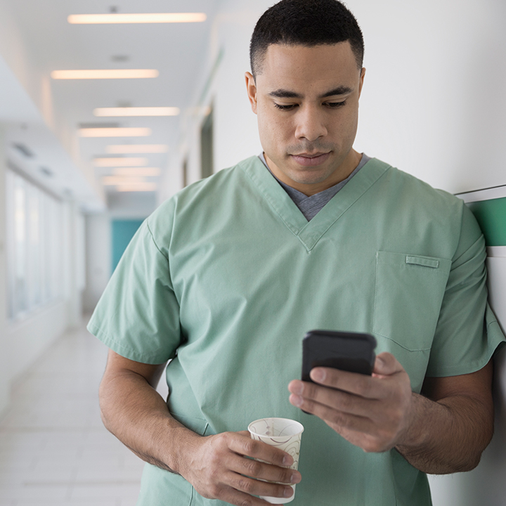Male nurse leaning against a wall holding a cup of water and his smartphone which he is sending messages through secure conversations with