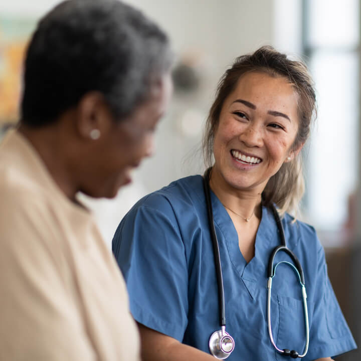 Female skilled nursing provider with a stethoscope around her neck seated and smiling with an elderly female resident