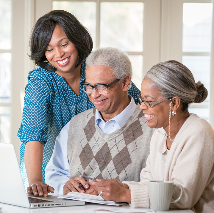 Female home health care provider meeting and laughing with an elderly couple in their home reviewing information on a laptop
