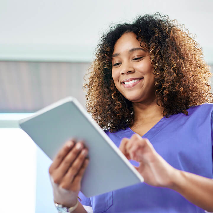 Female skilled nursing provider in purple scrubs smiles while looking at patient data in PointClickCare's software on a tablet device