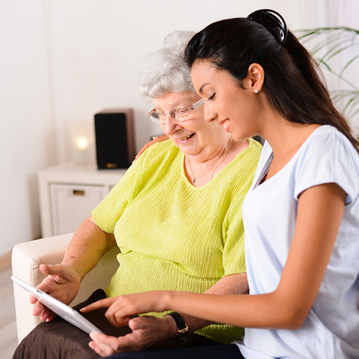Female home health care provider seated with an elderly female resident in her home reviewing information on a tablet device