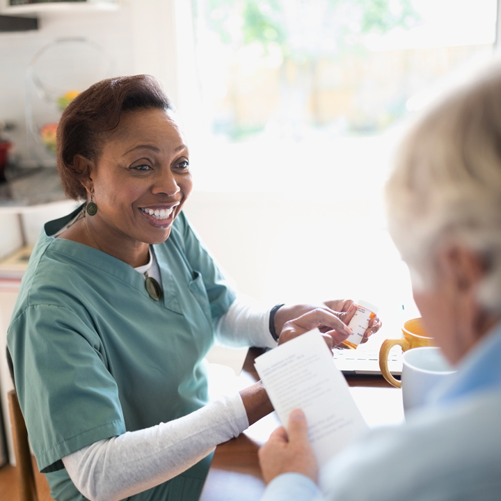 Female home health care provider in scrubs sitting with a resident in their home and reviewing prescription medication with them