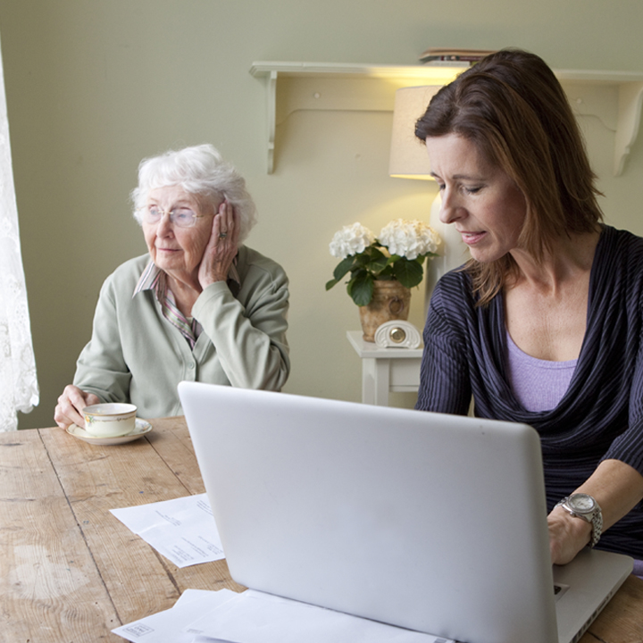 Female home health care provider seated going on patient documents and typing on a laptop while the patient drinks a cup of tea