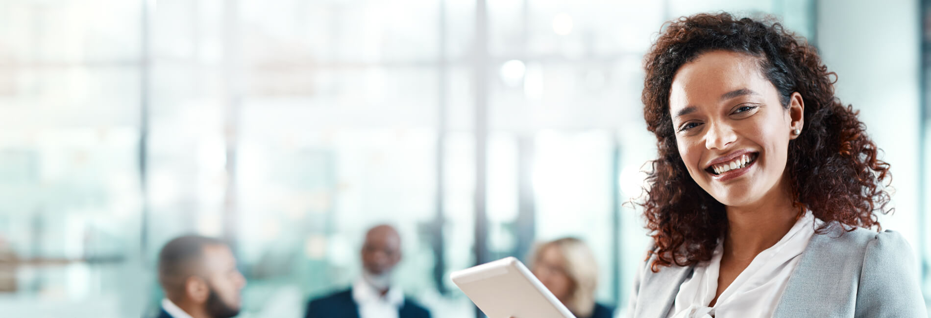 Female business executive standing and smiling with a tablet device with professionals seated in the background