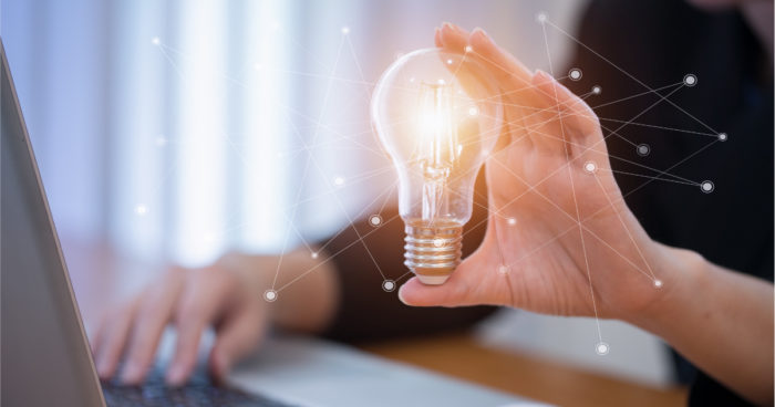 A photo of a person holding a lightbulb while also using PointClickCare software on a laptop.