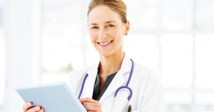 A female physician smiles for the camera while using PointClickCare software on a tablet.