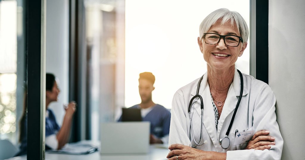 A doctor leaning against a wall and an out of focus nurse sitting at a table in the background