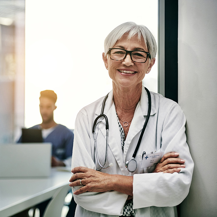 A doctor leaning against a wall and an out of focus nurse sitting at a table in the background