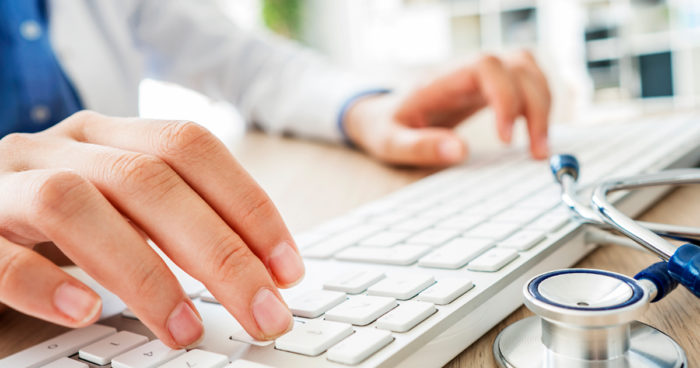 A close-up image of a doctor's hands typing on a computer keyboard.
