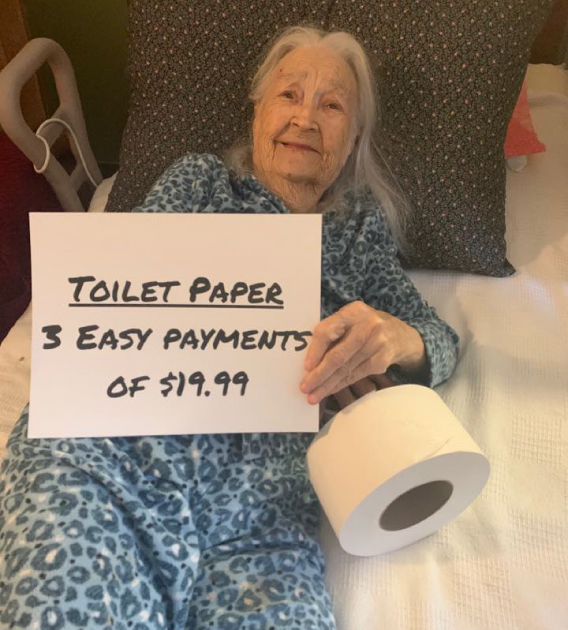 Elderly female resident at long-term care facility Silver Bluff Village holding a paper sign and with a toilet paper roll next to her during COVID-19