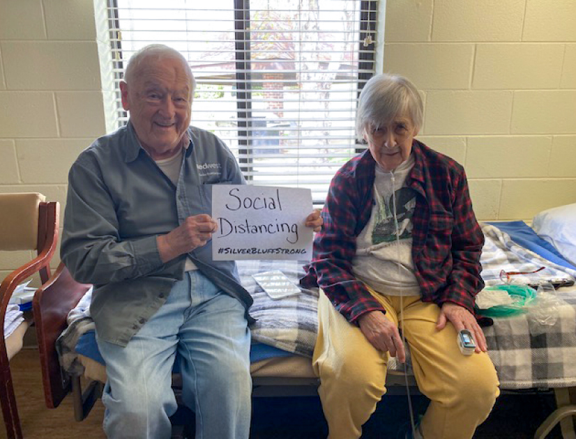 Elderly female and male long term care residents at Silver Bluff Village holding a social distancing paper sign during COVID-19 pandemic