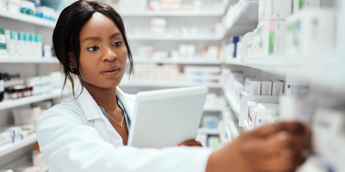 Female pharmacist using a medication management software on a tablet device as review pharmaceutical medications on the shelves