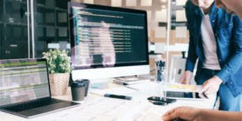 Two UX designers around a desk that has a laptop, desktop, tablet and paper on it