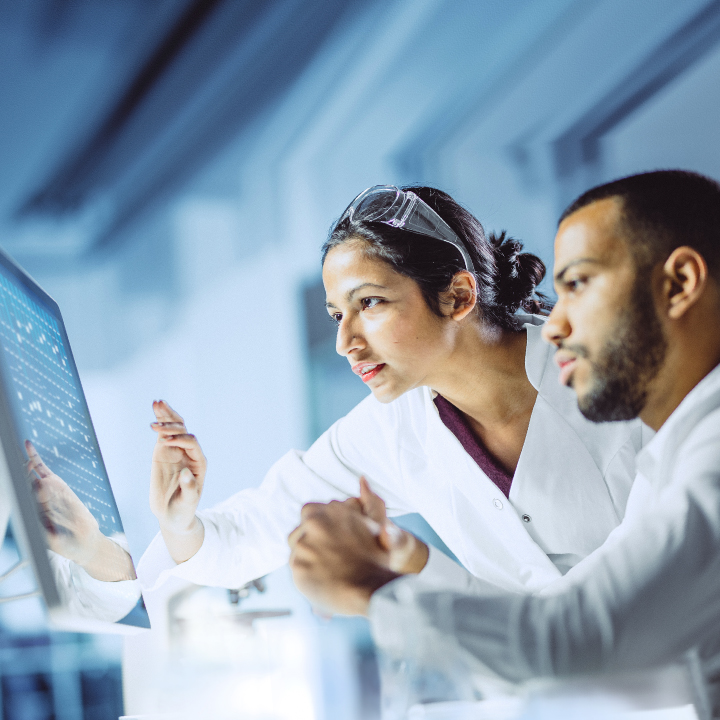 Male and female engineers in white lab coats reviewing data together on a large monitor