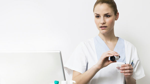 Female skilled nursing provider standing with prescription medication in her hand as she looks at eMAR data on a desktop monitor
