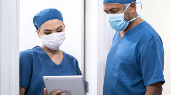 Two skilled nursing providers wearing scrubs and masks reviewing data on a tablet device together