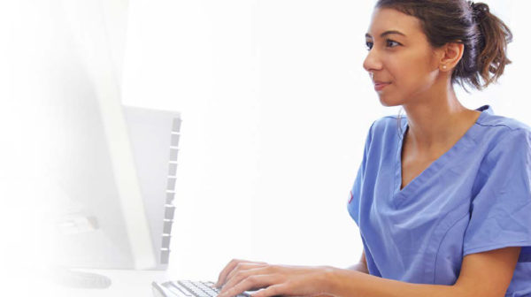 Female skilled nursing provider in purple scrubs seated and working on a desktop computer
