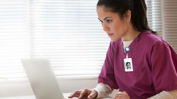Female skilled nursing provider seated in purple scrubs and working on a laptop