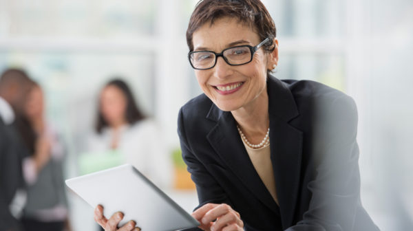 Female skilled nursing executive standing and smiling as she leans over while holding a tablet device