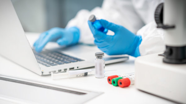 Close up of a lab technician's hands with gloves on using a laptop and holding a vile with medication