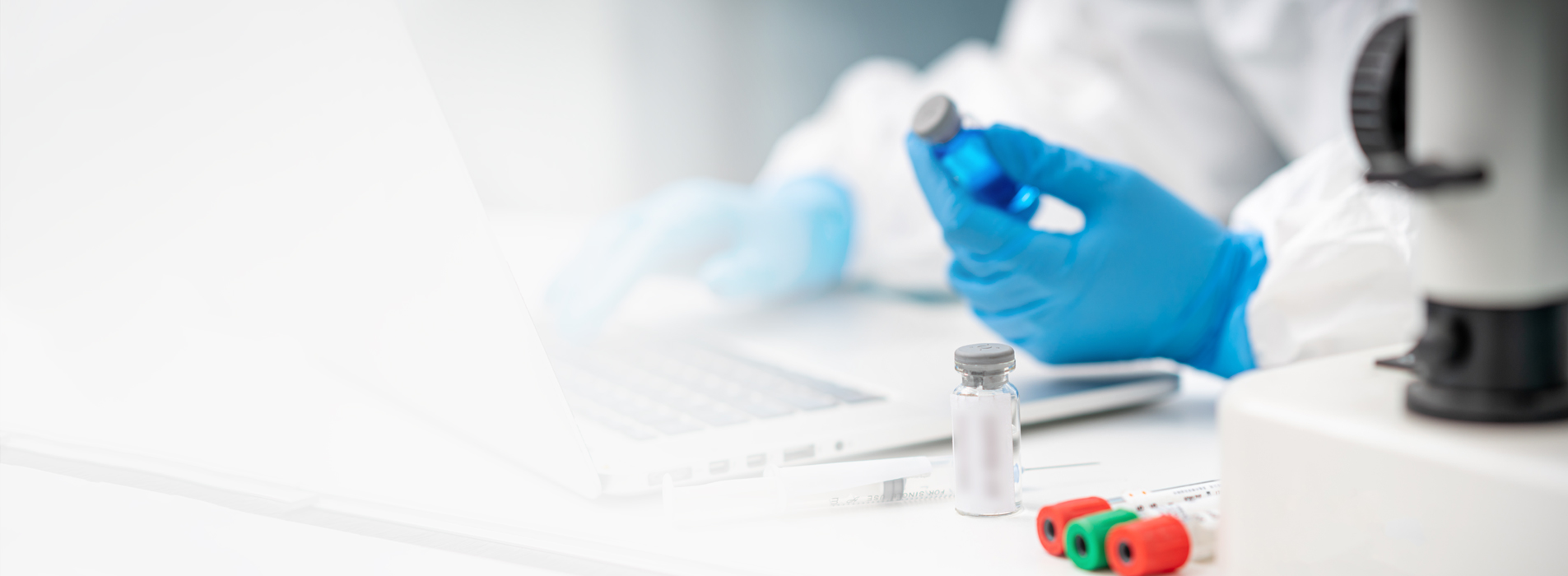 Close up of a lab technician's hands with gloves on using a laptop and holding a vile with medication