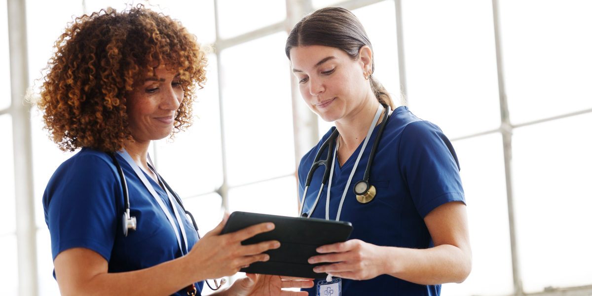 Two female skilled nursing providers with stethoscopes around their necks holding a tablet device together and looking at data