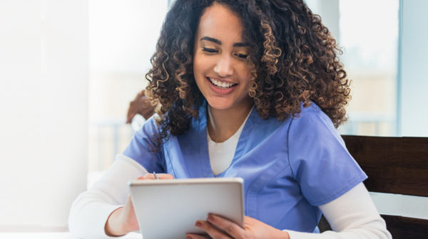 Female skilled nursing provider in scrubs sitting and smiling as she looks down at a tablet device using Automated Care Messaging