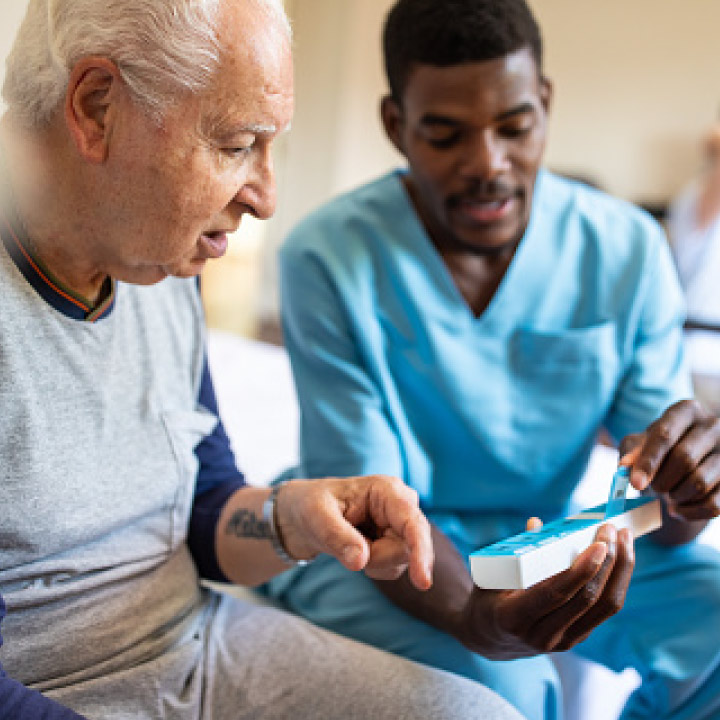 Male senior living care provider sitting with a male resident and showing him a weekly medication organizer