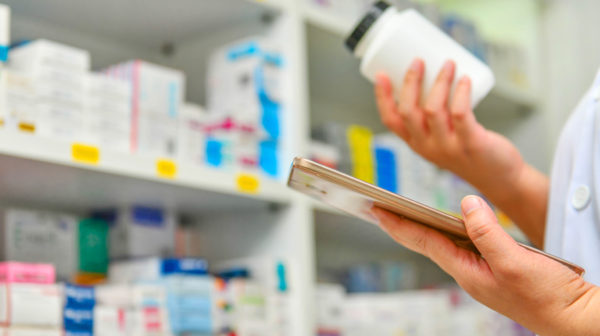 Close up of a pharmacist's hands in a pharmacy setting with one holding a tablet device and the other review a prescription medication bottle