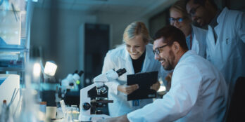Team of four scientists in a lab with no lights looking and smiling at something they see using a microscope