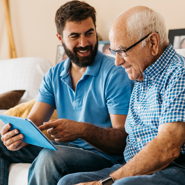 Male assisted living provider seated next to a male resident and both smiling as they review documentation on a tablet device