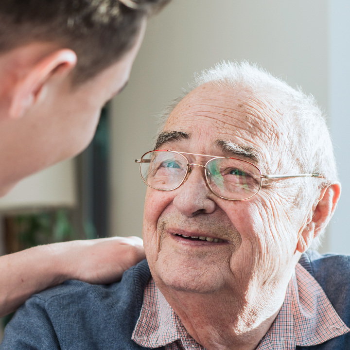 Close up of a male senior living resident smiling as a male senior living care provider puts his hand on his shoulder and speaks with him