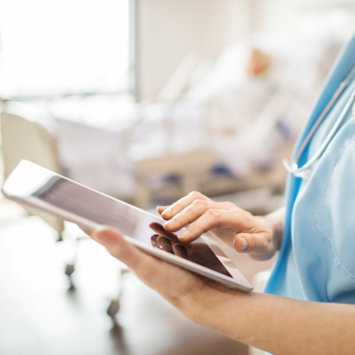 Close up of an acute care nurse's hands holding and using a tablet device with a patient in the background blurred