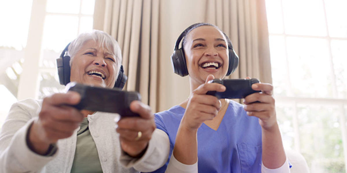 Female skilled nursing provider and resident sitting next to each other and laughing while they play video games with headsets on