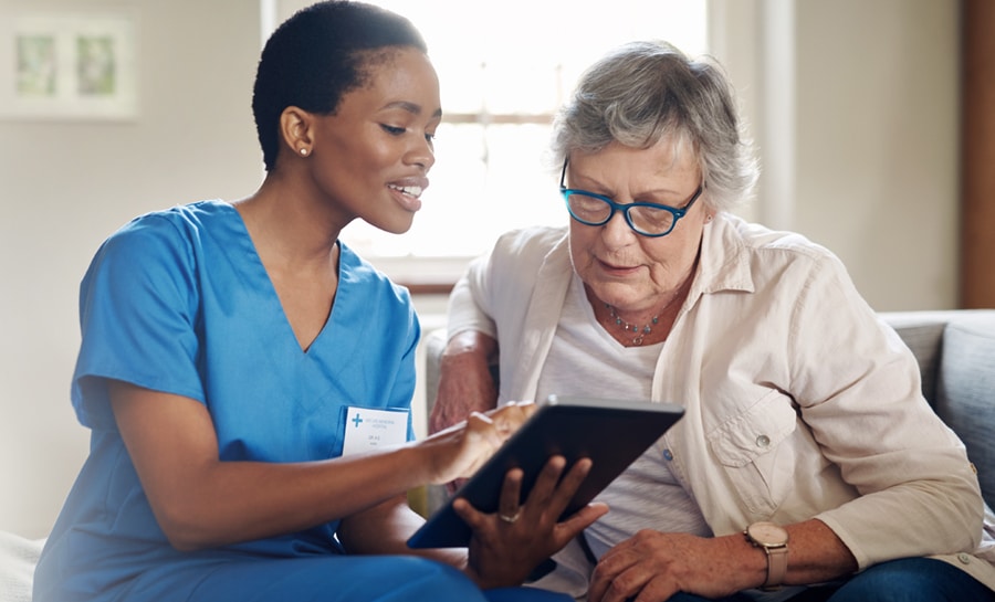 Female nurse in scrubs sitting with an elderly female patient and smiling as they review information on a tablet device together