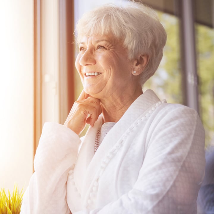 Elderly female resident standing in a white rob next to windows and smiling with one hand on her face