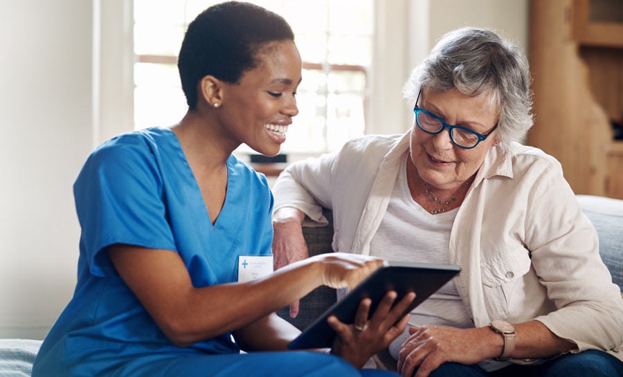 Female nurse in scrubs sitting with an elderly female patient and smiling as they review information on a tablet device together