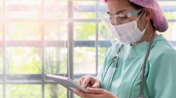 Female skilled nursing provider wearing scrubs, hair cover, face mask and face shield standing close to a window using a tablet device