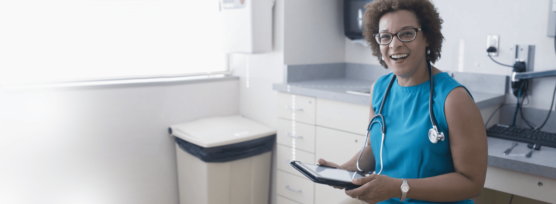 Female physician with a stethoscope around her neck standing in her office and smiling and she holds a tablet device