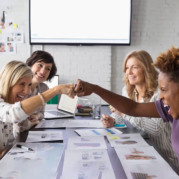 Group of female business professionals sitting around a desk smiling with two of them giving each other fist bumps