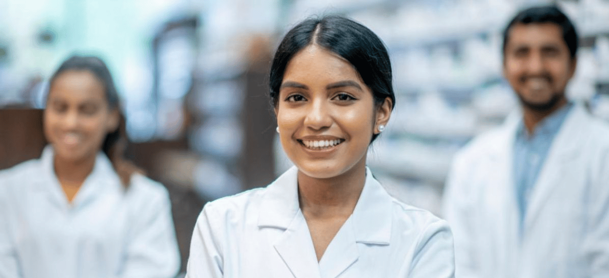 Close up of a female pharmacist standing and smiling in a pharmacy with two pharmacists standing behind blurred in the background