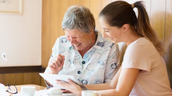 Female assisted living care provider sitting with a female resident and laughing as they review something on a tablet device