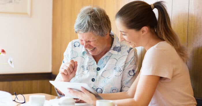 Female assisted living care provider sitting with a female resident and laughing as they review something on a tablet device