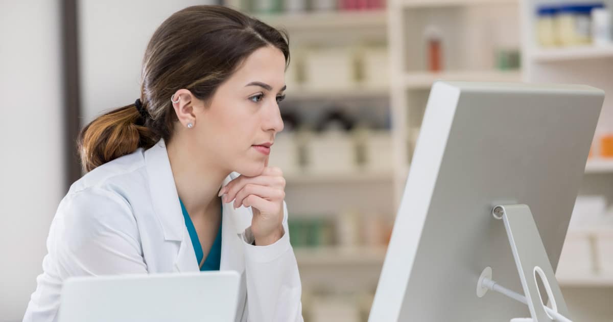 A female pharmacist working in a long-term care pharmacy using a desktop computer next to shelves of pharmaceutical medication