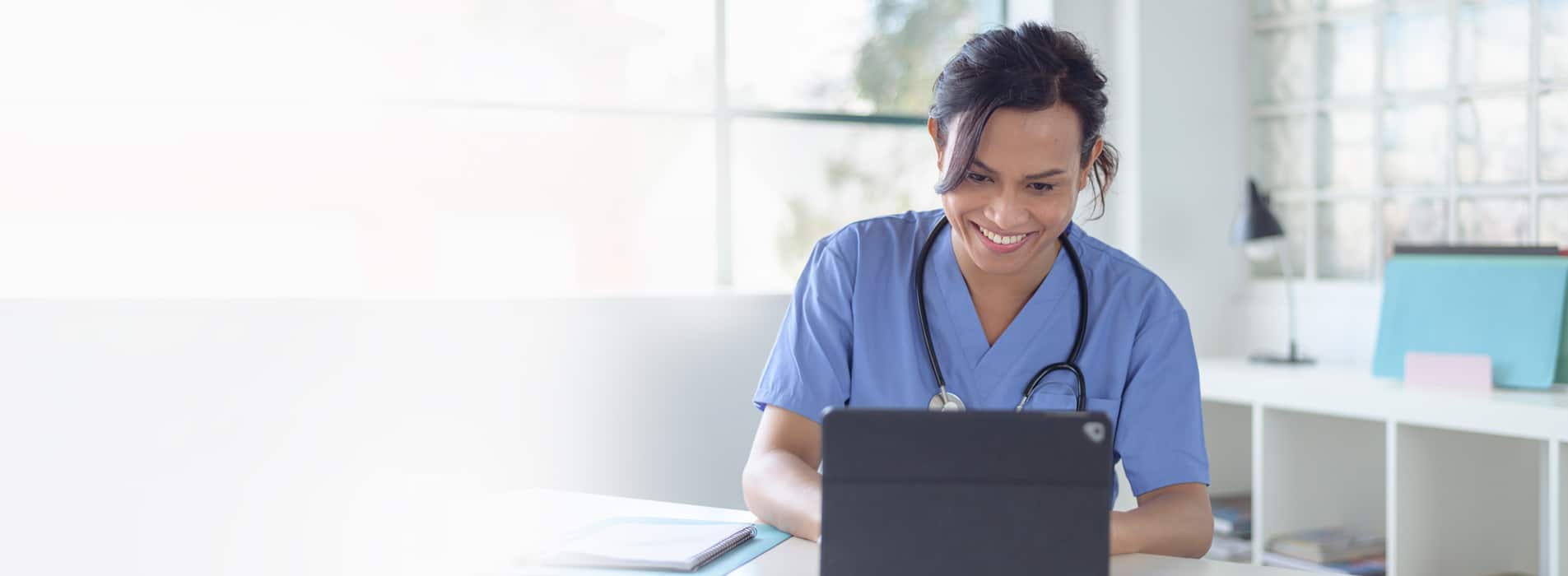 Female physician with scrubs on and a stethoscope around her neck provides virtual health services to a patient using a tablet device