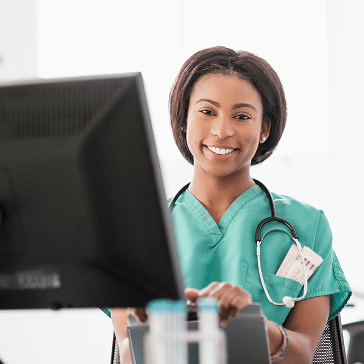 Female physician in green scrubs and with a stethoscope around her neck seated in front of a desktop computer and smiling