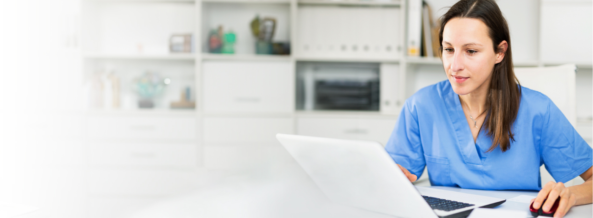 Photo of a female nurse with brown hair wearing blue medical scrubs working on her laptop