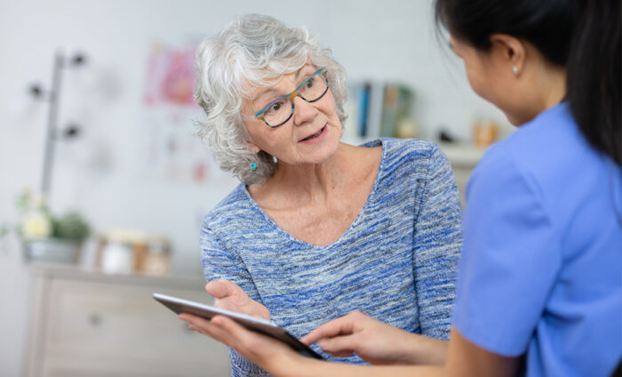 An elderly woman is speaking to her nurse as they review information on a tablet computer