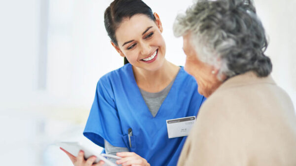 A female nurse is holding a medical chart and smiling while conversing with an elderly resident